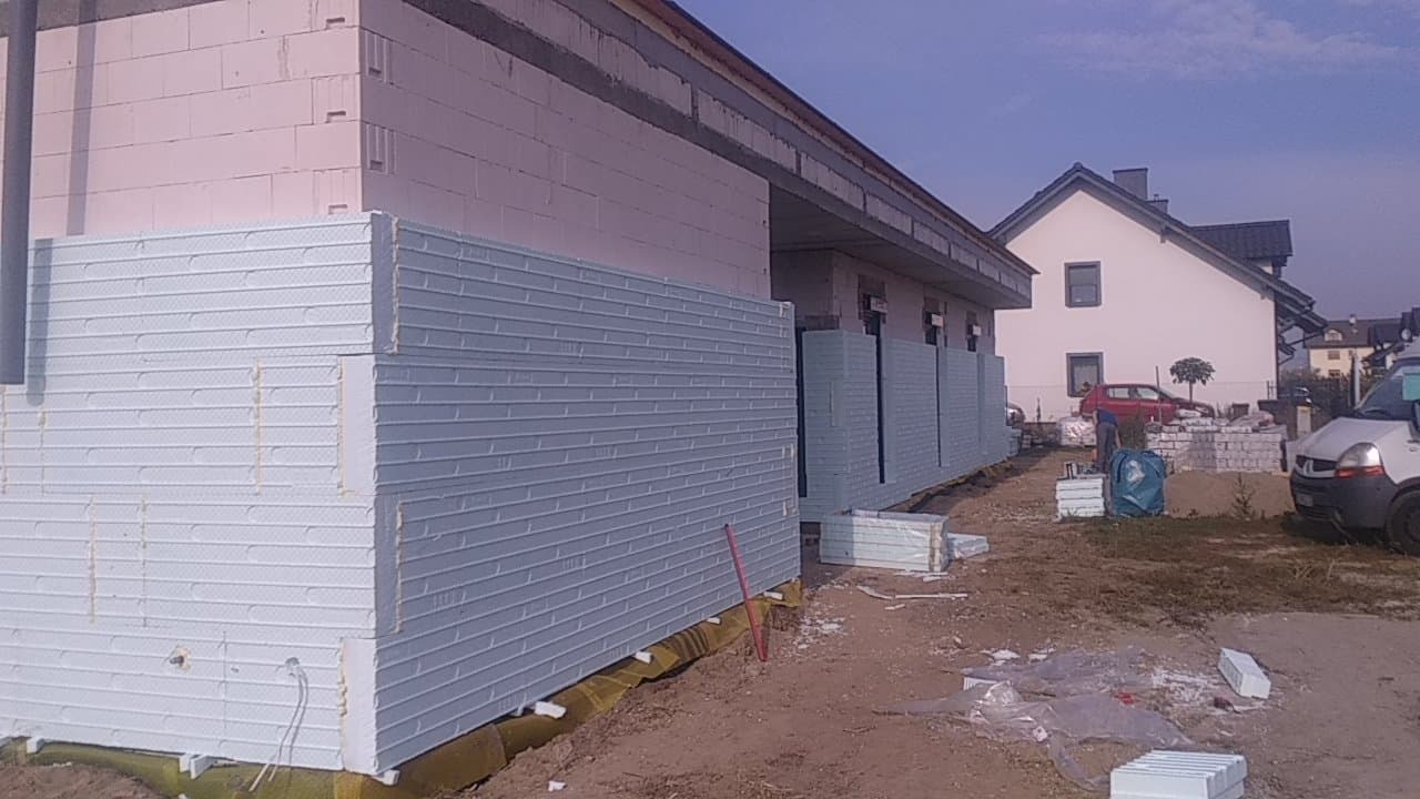 Budynek w trakcie prac izolacyjnych z widocznymi płytami styropianowymi, przygotowanymi do montażu na elewacji, na tle drugiego budynku i samochodu dostawczego.