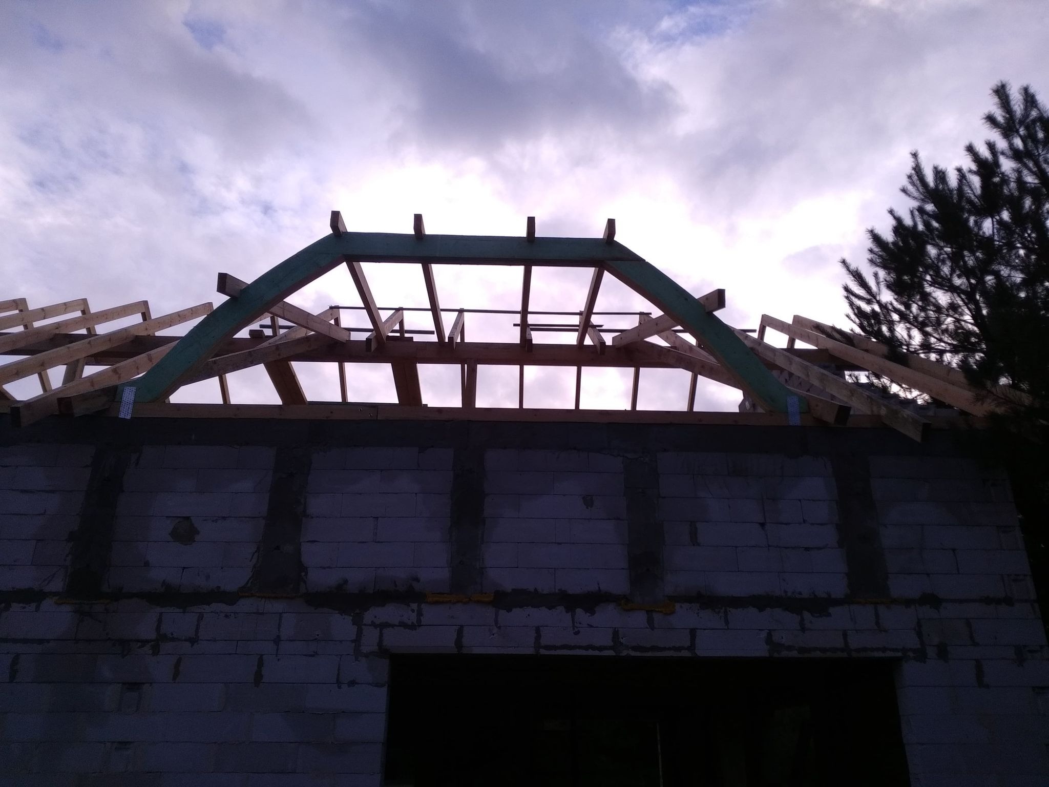 Wznoszenie dachu z drewnianą konstrukcją na murowanym budynku z bloczków, widoczne niebo i fragment drzewa.