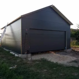 Nowoczesny, grafitowy garaż blaszany z bramą segmentową, widoczna drabina oparta o ścianę boczną, teren wokół w trakcie prac wykończeniowych, z piaskiem i narzędziami.