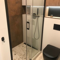 ŁazienkiSV - Nowoczesna łazienka z kabiną prysznicową typu walk-in z czarnymi ramami, toaleta w kolorze czarnym, płytki podłogowe lastryko, ściana z efektem rdzy i drewniana półka z podświetleniem LED.