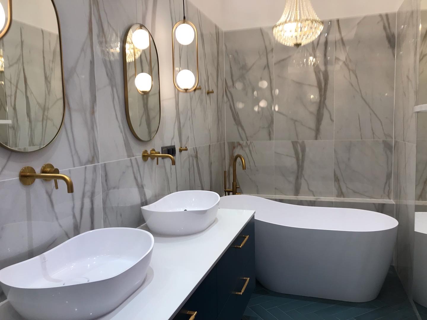 Elegancka łazienka z marmurowymi płytkami, dwiema owalnymi umywalkami nablatowymi, złotą armaturą, wiszącymi lampami i kryształowym żyrandolem nad wolnostojącą wanną.
