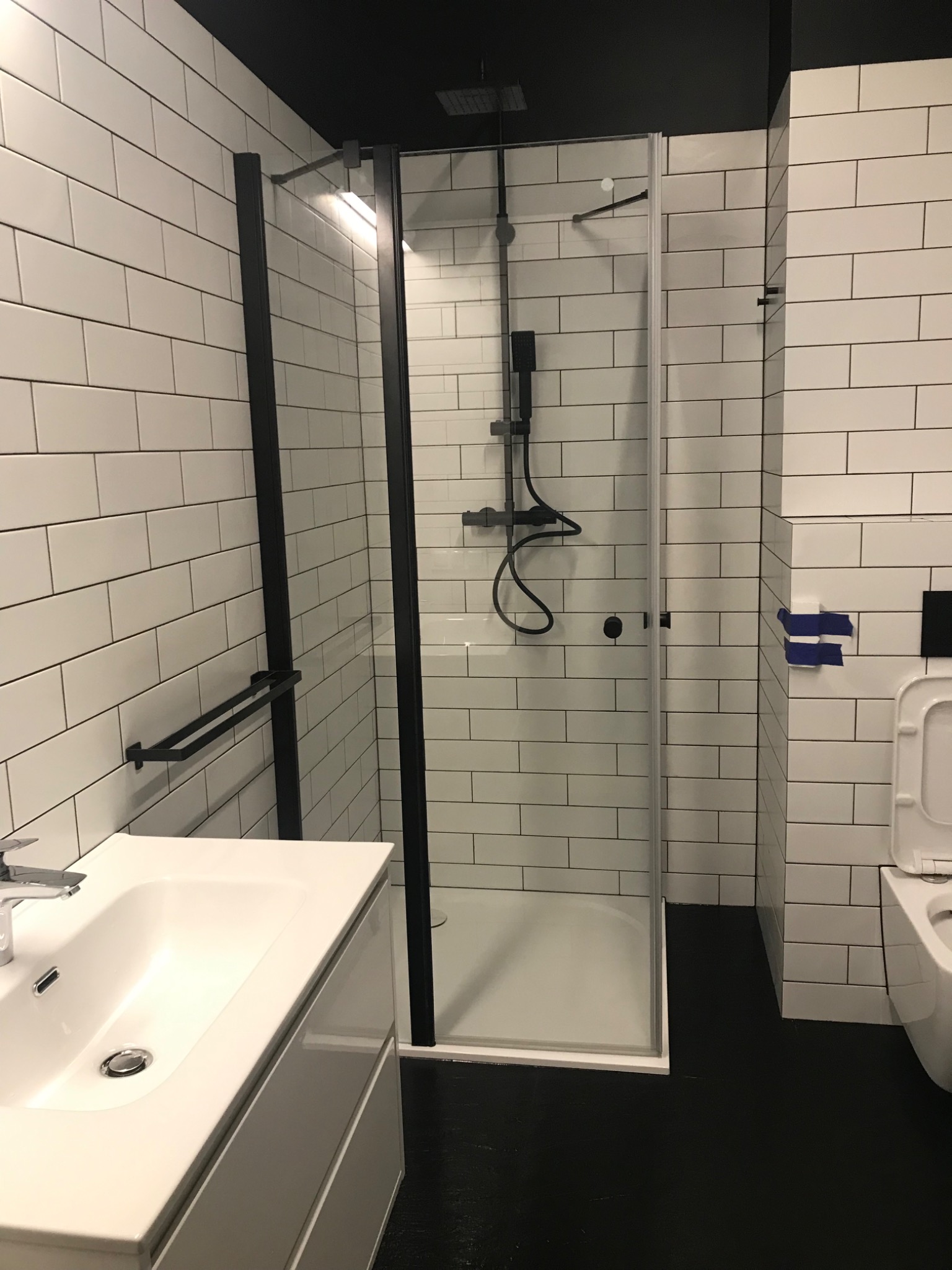 Nowoczesna łazienka z białymi, prostokątnymi płytkami, czarną armaturą i kabiną prysznicową typu walk-in, widoczna umywalka z szafką, czarna podłoga i sufit, niebieska taśma malarska na ścianie.