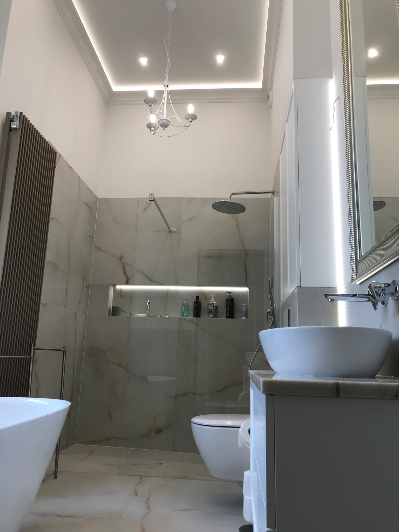 Nowoczesna łazienka z marmurowymi płytkami, okrągłą umywalką nablatową, kabiną prysznicową typu walk-in i grzejnikiem drabinkowym w kolorze miedzi.