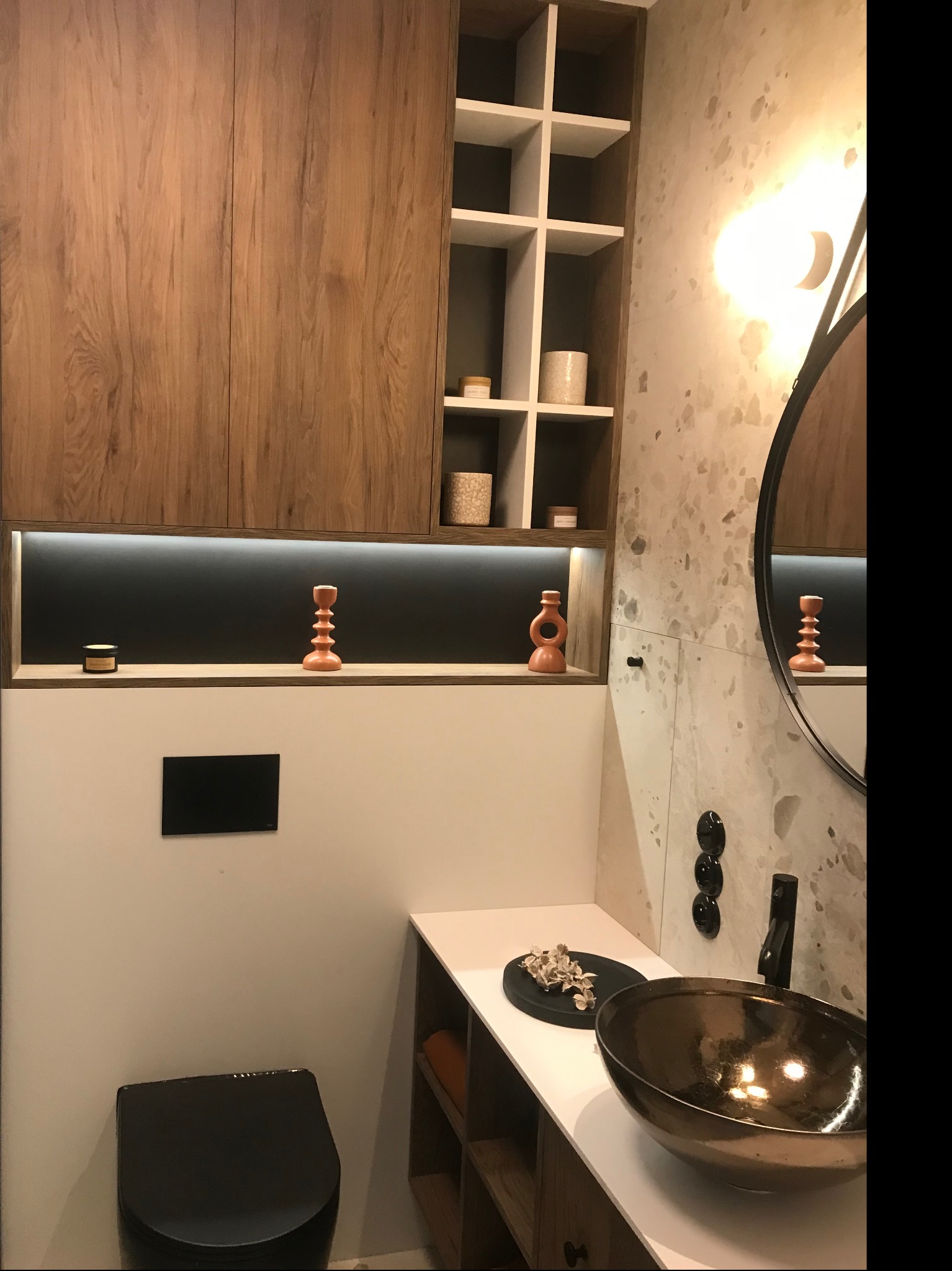 Nowoczesna łazienka z drewnianymi szafkami, białą umywalką w kolorze brązu, czarną toaletą i dekoracyjnymi elementami oświetleniowymi.