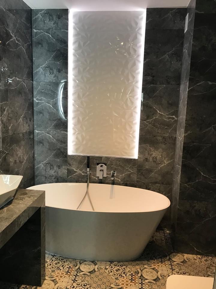 Nowoczesna łazienka z wolnostojącą wanną, szarymi marmurkowymi płytkami ściennymi i podświetlaną kolumną z białych płytek 3D, wzorzysta podłoga.