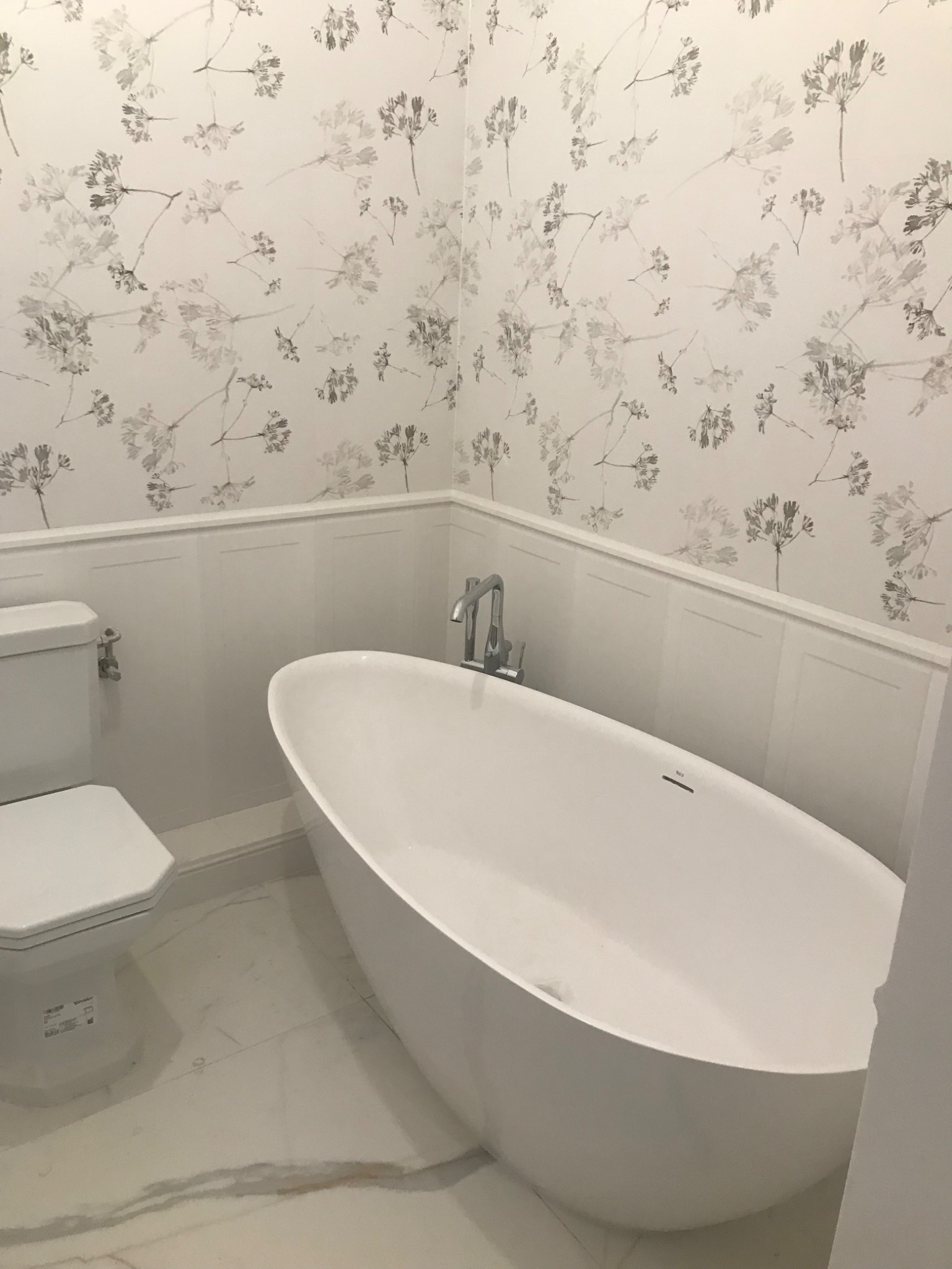 Nowoczesna biała wanna wolnostojąca z chromowaną baterią, biała toaleta oraz ściany wykończone w białej boazerii i tapetą w delikatny kwiatowy wzór na marmurowej podłodze.