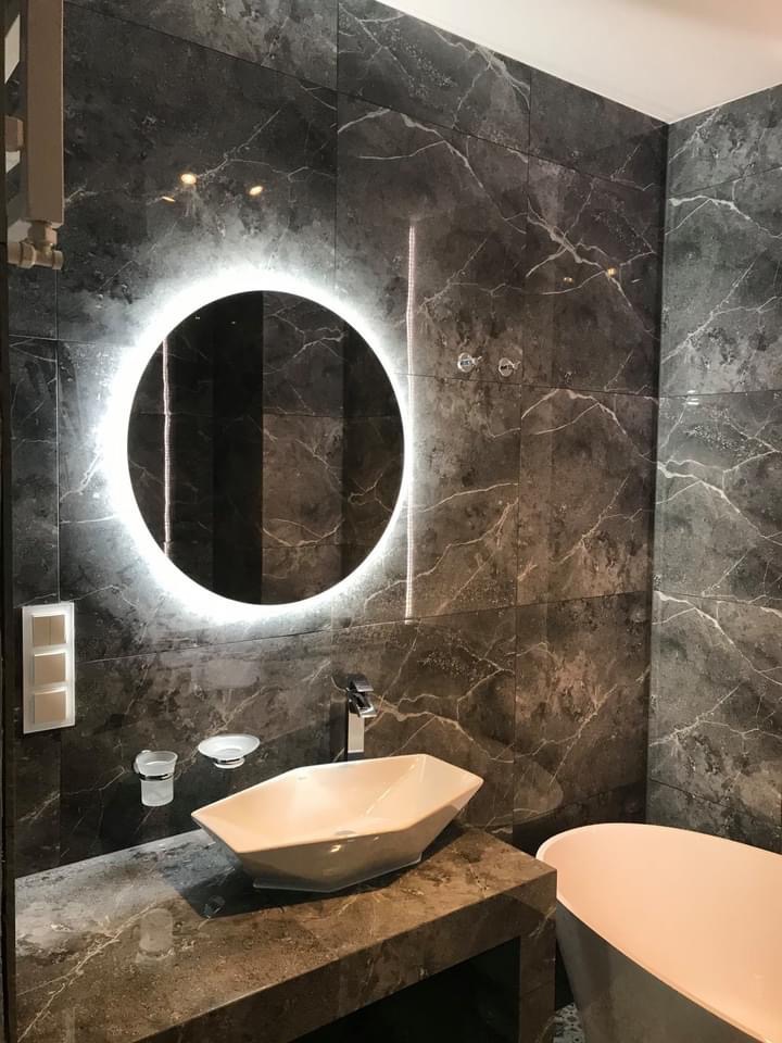 Elegancka łazienka w ciemnych marmurkowych płytkach z okrągłym podświetlanym lustrem, designerską umywalką nablatową i minimalistyczną baterią.