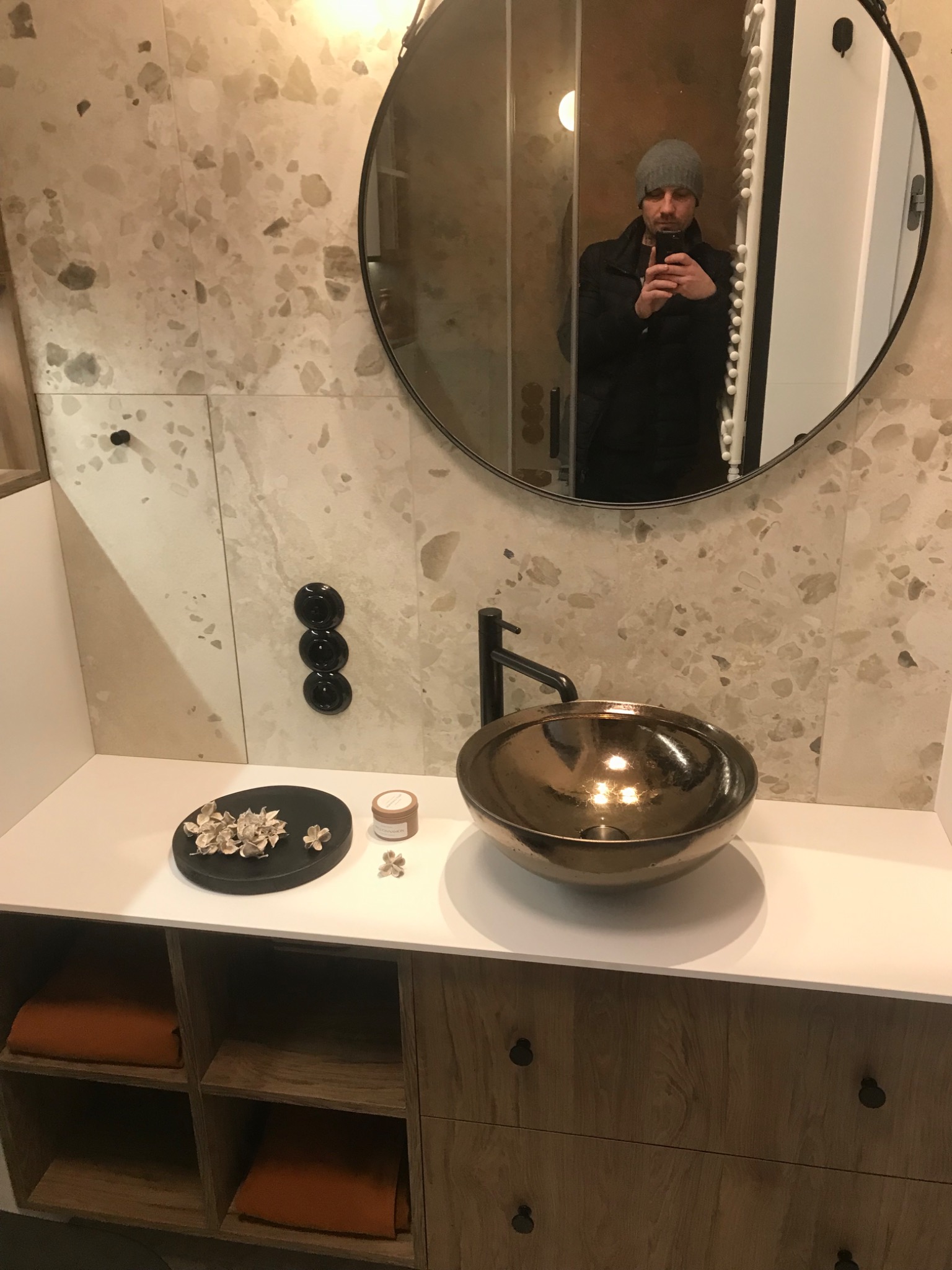 Nowoczesna łazienka z okrągłym lustrem, brązową umywalką nablatową i armaturą w kolorze czarnym, dekoracje w postaci suszonych kwiatów na blacie.