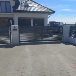 Nowoczesna, pozioma brama wjazdowa z szarego metalu i jasnymi słupkami, na tle domu z ciemną dachówką i podjazdu z kostki brukowej, częściowo widoczny samochód dostawczy.