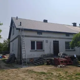 Dom jednorodzinny z nowym, szarym dachem z blachy trapezowej, drabina oparta o ścianę, widoczne kominy i obróbki blacharskie, teren wokół domu częściowo zagospodarowany.