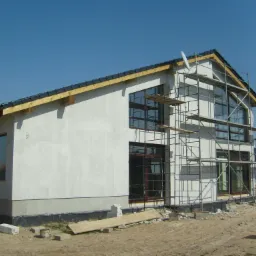 Budowa domu jednorodzinnego z białym tynkiem, widoczne rusztowania, okna z ciemnymi ramami i antena satelitarna na dachu, w tle kolejny dom w trakcie budowy.