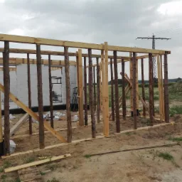 Wznoszenie szkieletu drewnianego domu z bali na fundamencie z bloczków, widoczne zbrojenie w narożniku, teren budowy z piaskiem i konstrukcją wsporczą, słupy energetyczne w tle.