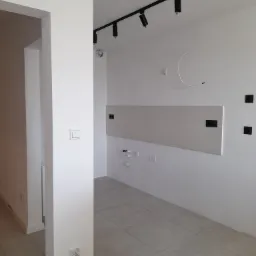 Wnętrze po remoncie w Wieliczce: świeżo położona jasna glazura na ścianie, szara podłoga z płytek, białe ściany z czarnymi gniazdkami i czarną listwą oświetleniową na suficie. Widoczny fragment...