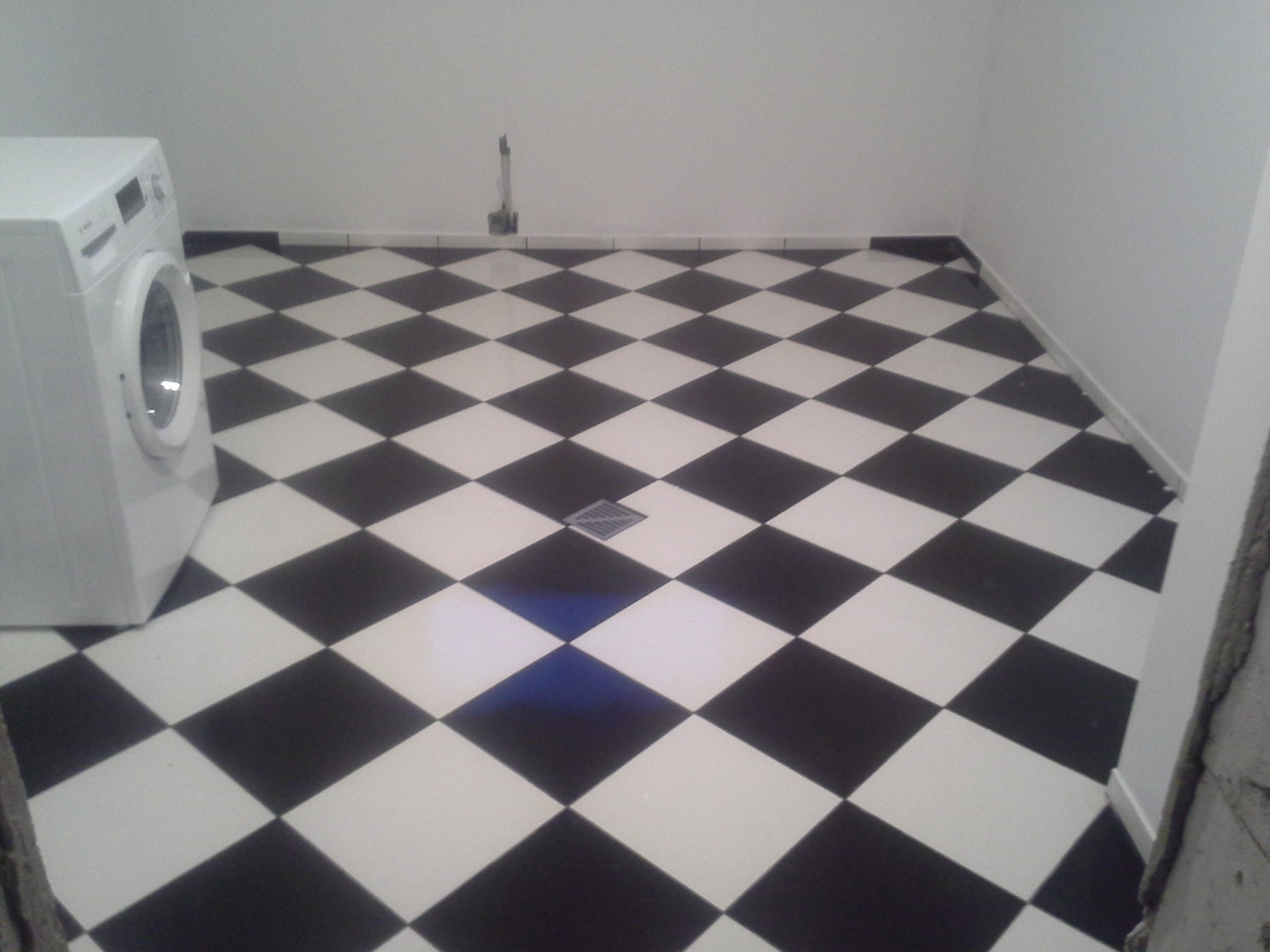 Ułożona pod kątem podłoga w łazience z czarno-białych płytek w szachownicę, widoczny odpływ liniowy i pralka z boku.