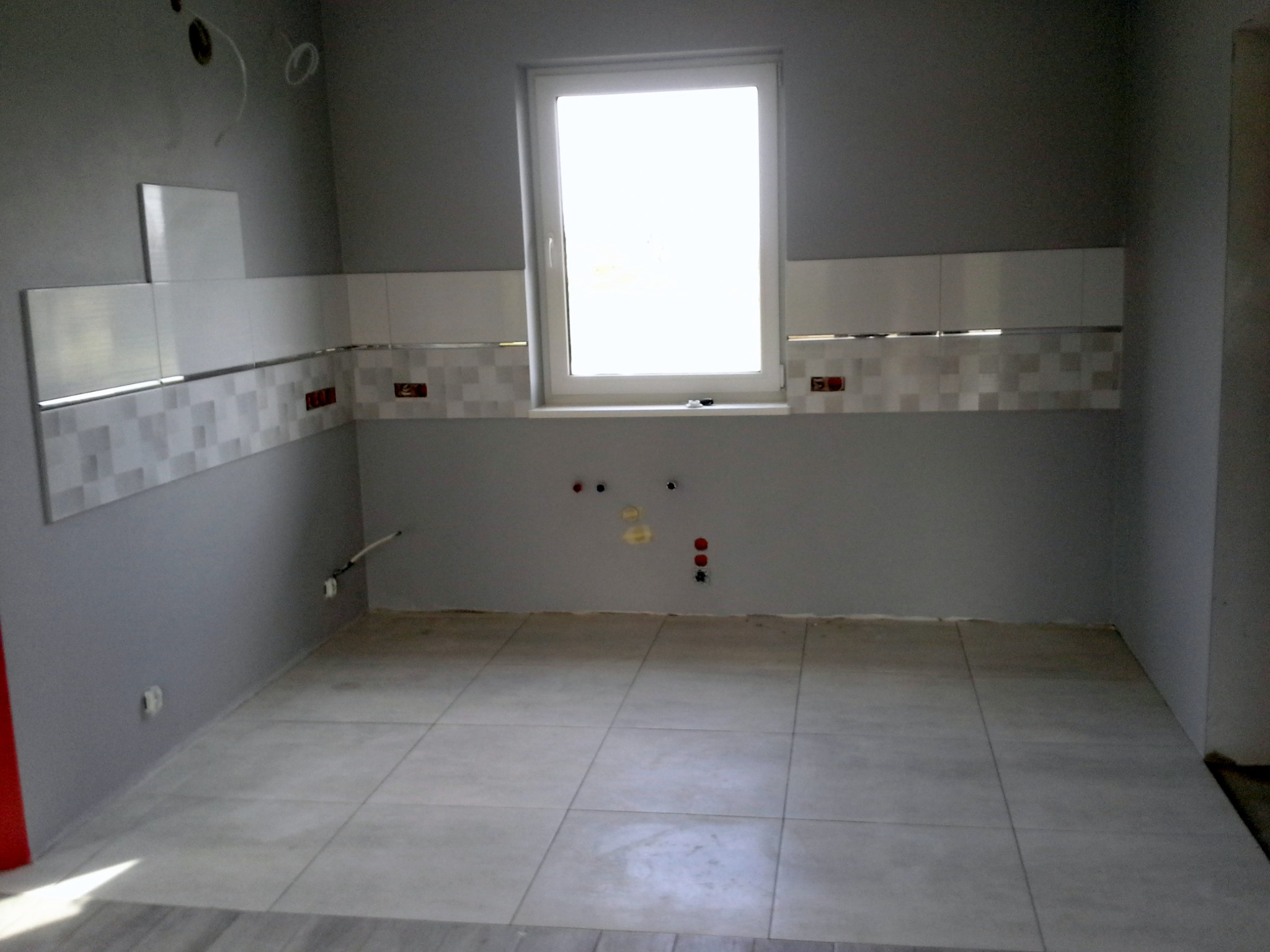 Wnętrze łazienki w trakcie wykańczania: ułożone jasne płytki podłogowe i częściowo położona glazura ścienna z dekoracyjnym pasem, widoczne instalacje elektryczne i hydrauliczne przed montażem...