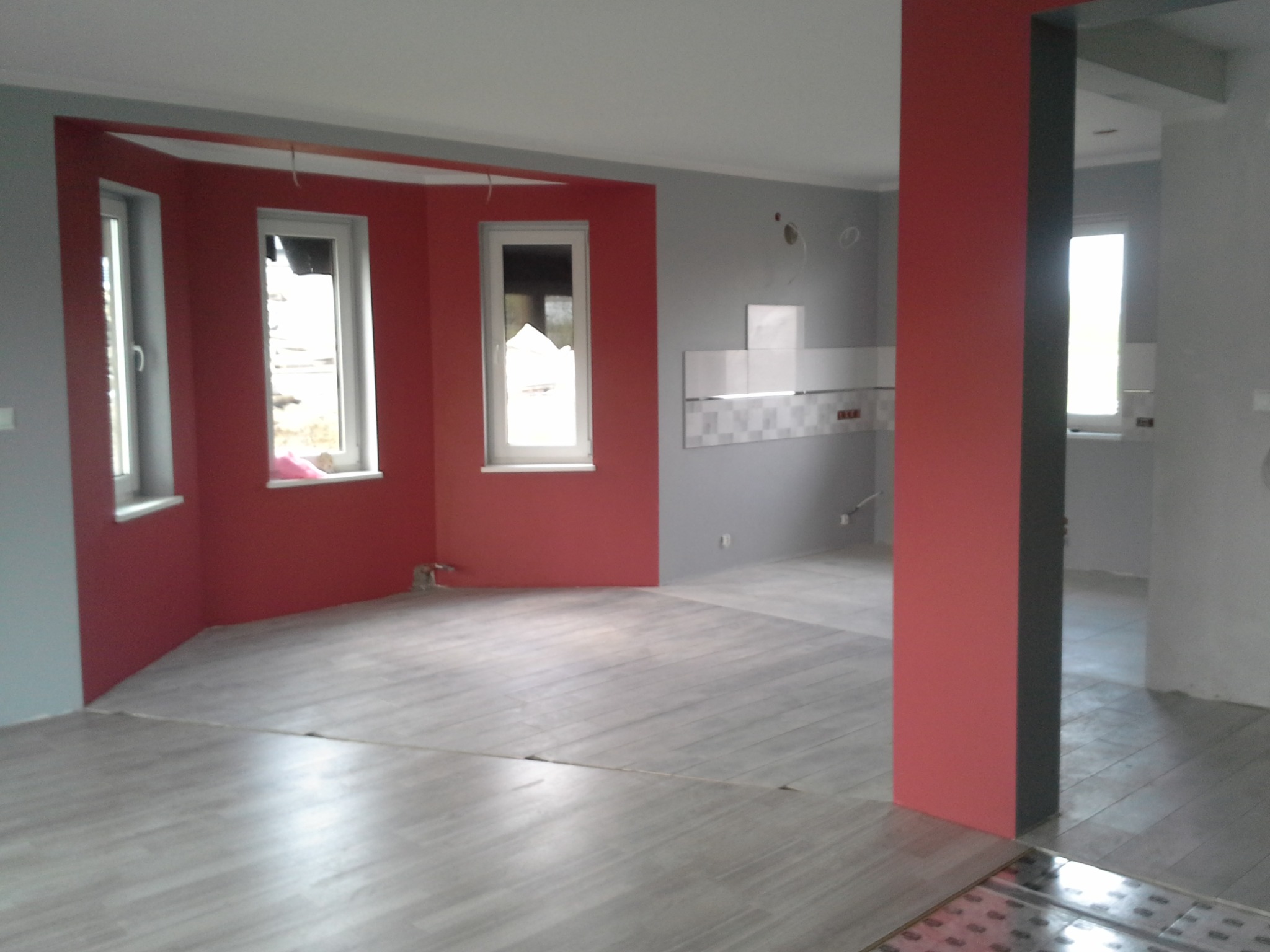 Pomalowane na szaro i czerwono wnętrze z ułożoną podłogą imitującą drewno, widoczne okna i fragment ściany z płytkami.