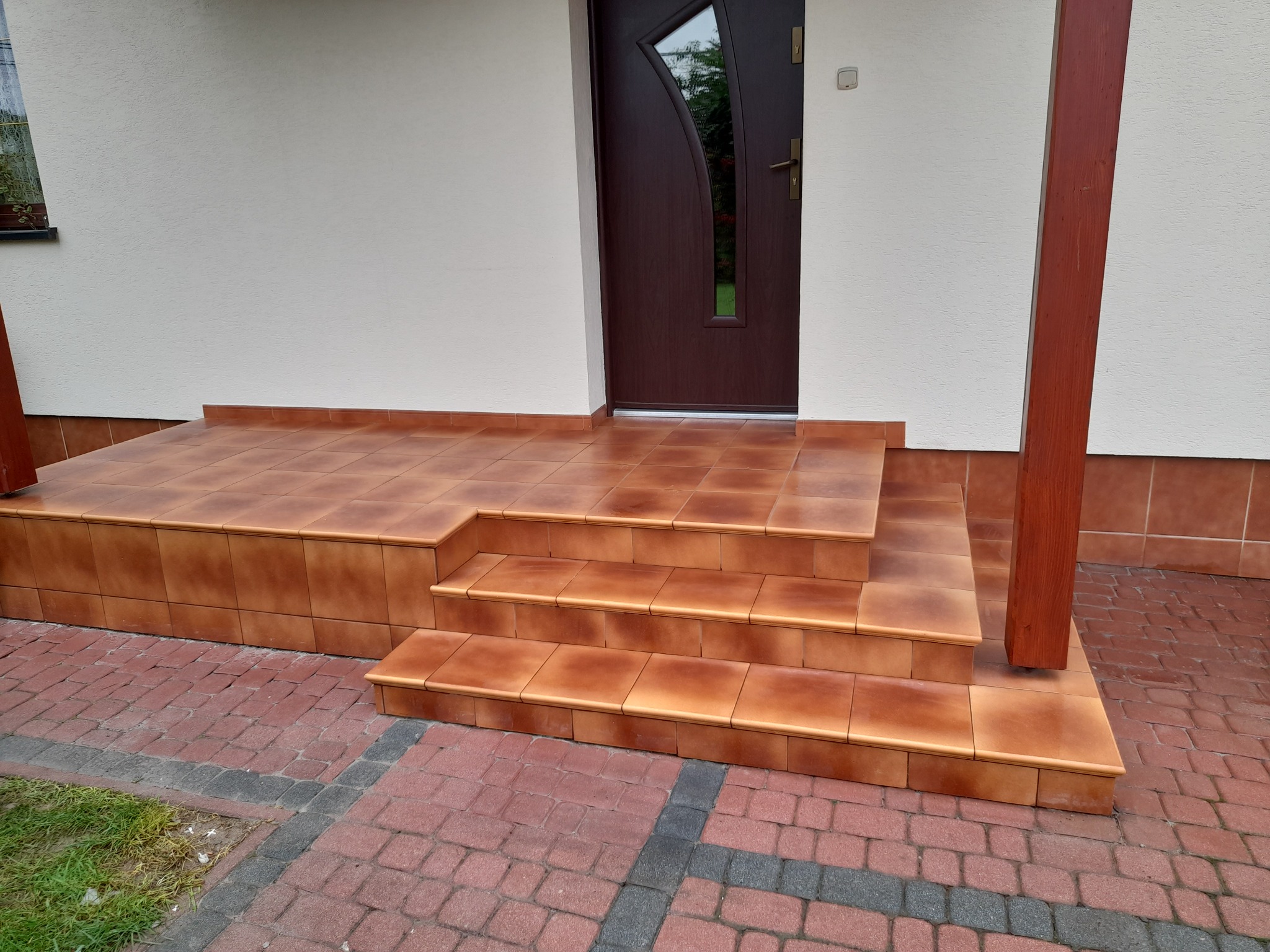 Kafelkowane schody wejściowe z brązowych płytek prowadzące do brązowych drzwi wejściowych, obok drewniana kolumna werandy i kostka brukowa na podjeździe.