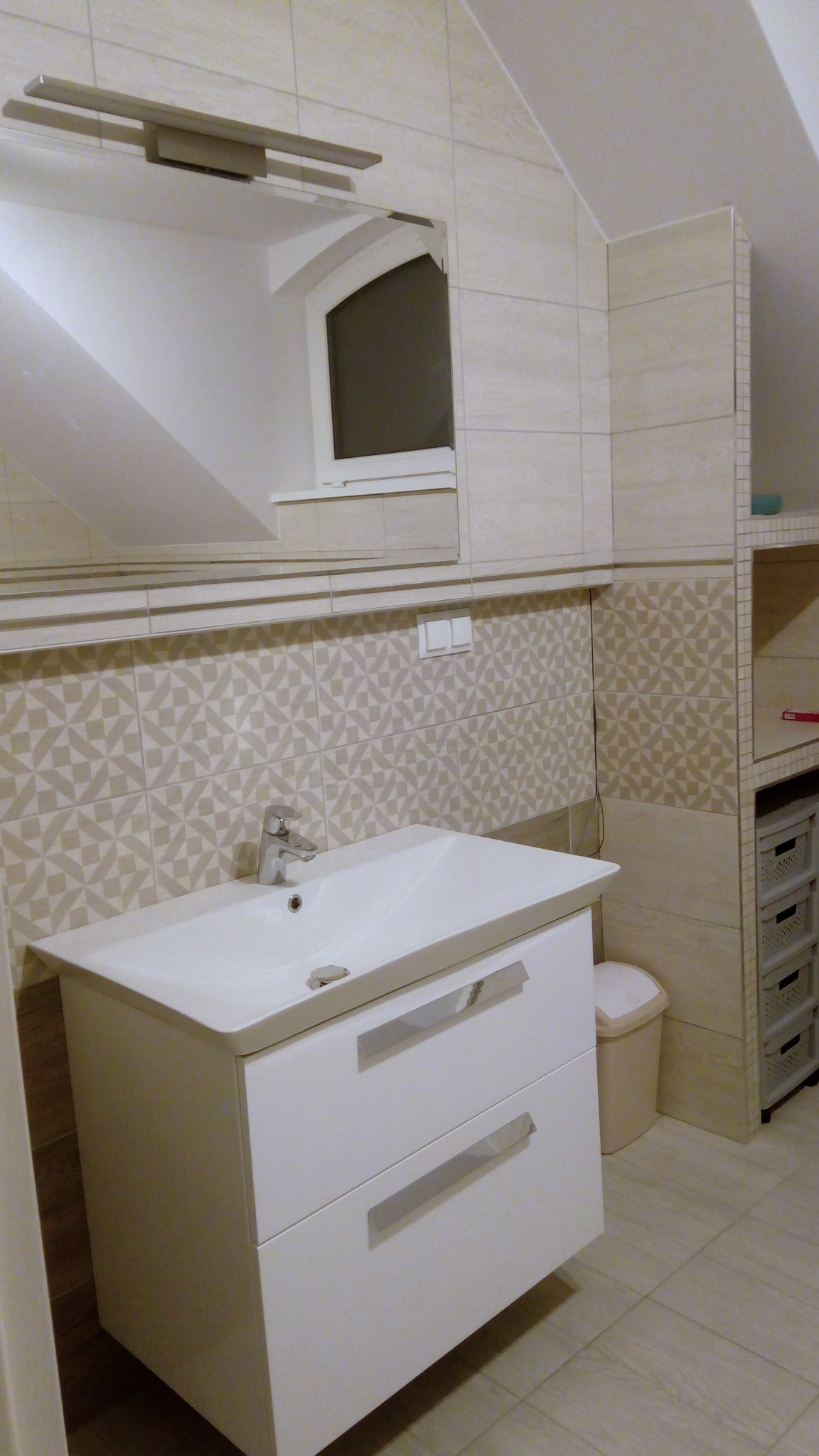 Umywalka z białą szafką z dwoma szufladami i chromowanymi uchwytami, bateria umywalkowa, wzorzyste płytki na ścianie, lustro z oświetleniem w łazience na poddaszu.