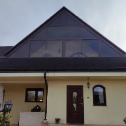 Dom z dużym trójkątnym oknem dachowym i brązową stolarką, widok z zewnątrz na elewację z beżowym tynkiem i schodami z piaskowca.