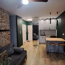 Salon z aneksem kuchennym po remoncie: ściana z cegły, szara sofa, drewniana podłoga, biała zabudowa kuchenna z wyspą i hokerami, ciemnozielone ściany.