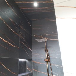 Waldek - Wykończona kabina prysznicowa z czarnymi płytkami imitującymi marmur z wyraźnym, złotym użyleniem, wbudowana półka z podświetleniem LED oraz deszczownica w kolorze złotym.