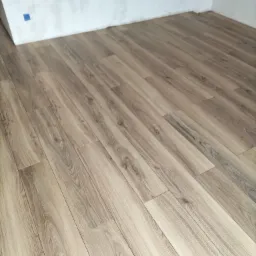 Jasne panele podłogowe z wzorem drewna ułożone w rogu pokoju w Wałbrzychu, ściany przygotowane do malowania.