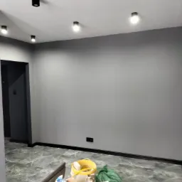 Świeżo pomalowane szare wnętrze z marmurową podłogą i zamontowanymi czarnymi lampami sufitowymi, z widocznymi narzędziami i materiałami budowlanymi w dolnej części kadru.