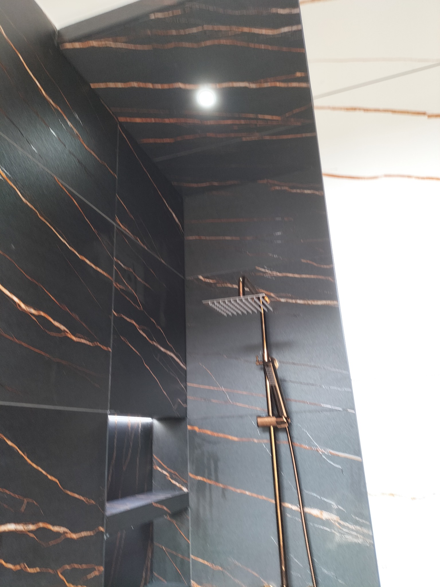 Wykończona kabina prysznicowa z czarnymi płytkami imitującymi marmur z wyraźnym, złotym użyleniem, wbudowana półka z podświetleniem LED oraz deszczownica w kolorze złotym.