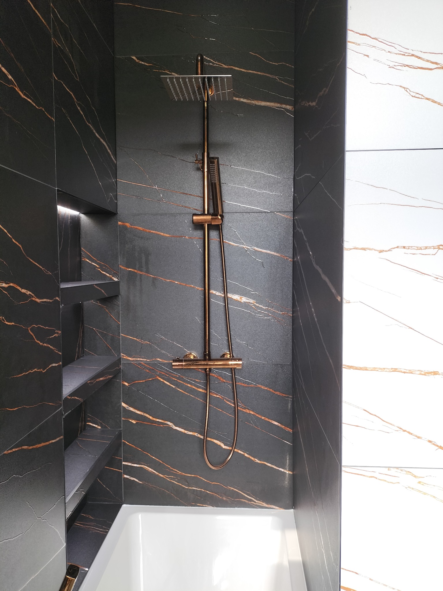Nowoczesna kabina prysznicowa z czarnymi płytkami imitującymi marmur z złotymi żyłami, półki wnękowe z podświetleniem LED, bateria i deszczownica w kolorze różowego złota, biała wanna.