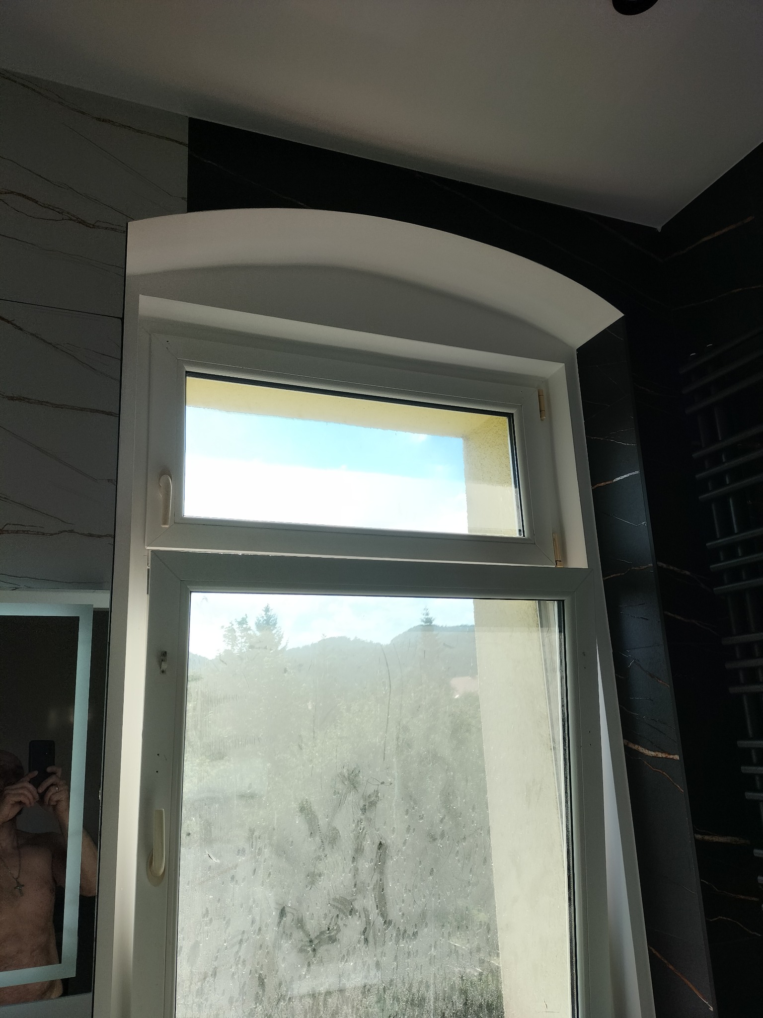 Okno łazienkowe z zaokrąglonym nadprożem, widok na zewnątrz z zamazaną szybą, obok czarne płytki imitujące marmur