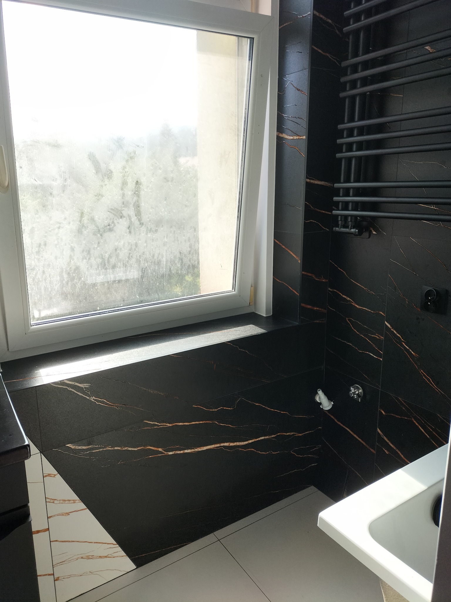 Nowoczesna łazienka z czarnymi płytkami imitującymi marmur ze złotymi żyłami, grzejnikiem drabinkowym w kolorze czarnym i białą ramą okienną, wykończona w minimalistycznym stylu.