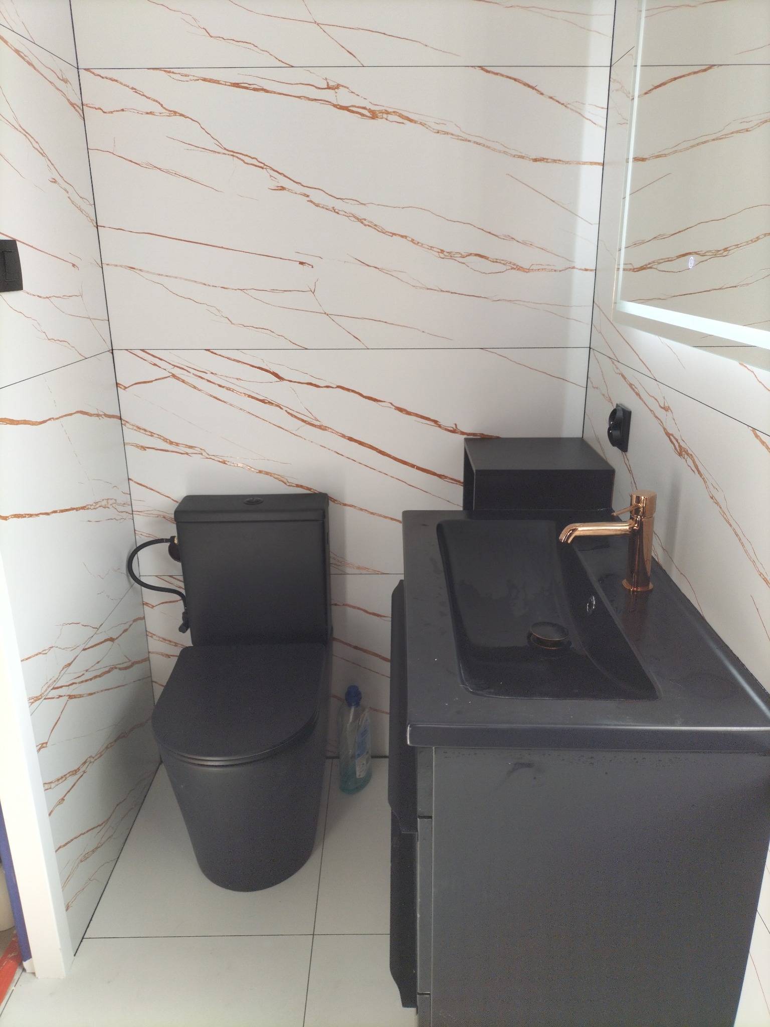 Nowoczesna łazienka z czarną armaturą i umywalką na szafce, białe płytki imitujące marmur z brązowymi żyłami, złota bateria umywalkowa.