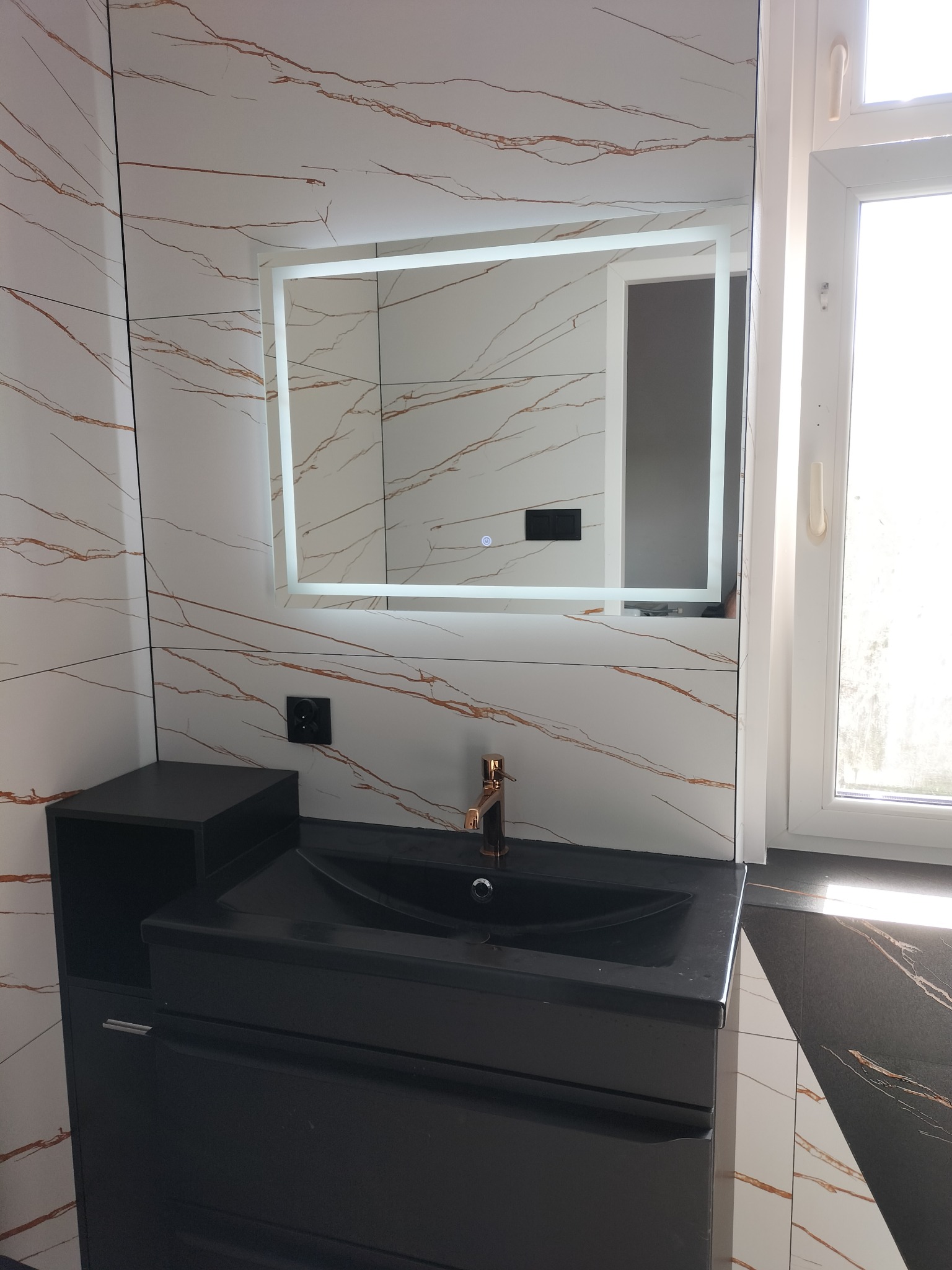 Czarna umywalka nablatowa z szafką, złota bateria, podświetlane lustro i marmurowe płytki z brązowymi żyłami w wykończonej łazience.