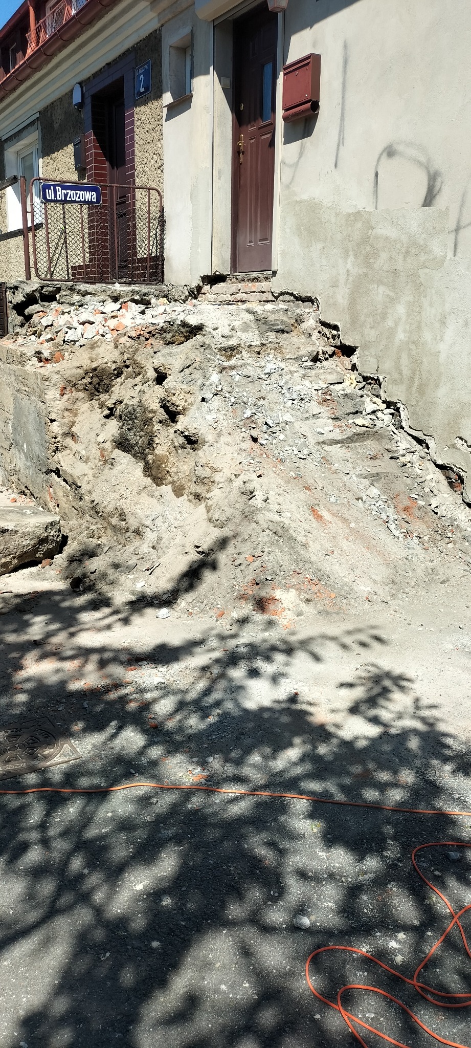 Demontaż schodów wejściowych do budynku z widocznymi fragmentami gruzu i cegieł, zarys budynku z drzwiami i skrzynką pocztową, cień drzewa na chodniku oraz pomarańczowy kabel na pierwszym planie.