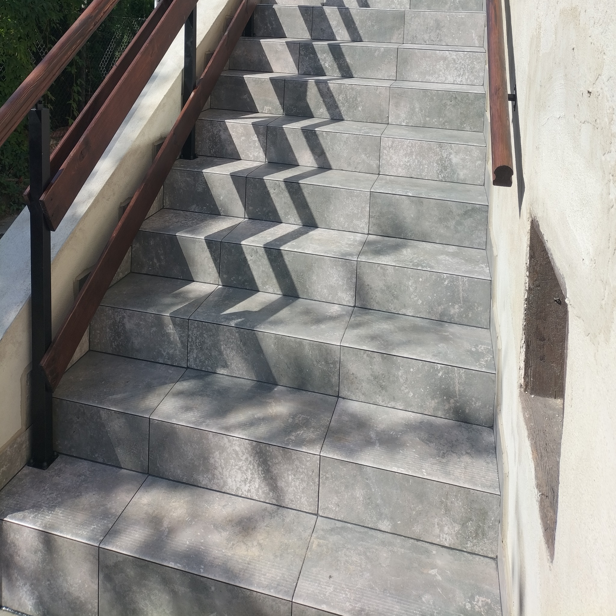 Zewnętrzne schody wyłożone szarą, kwadratową płytką ceramiczną z drewnianą balustradą po lewej stronie, oświetlone promieniami słonecznymi tworzącymi cienie na powierzchni płytek.