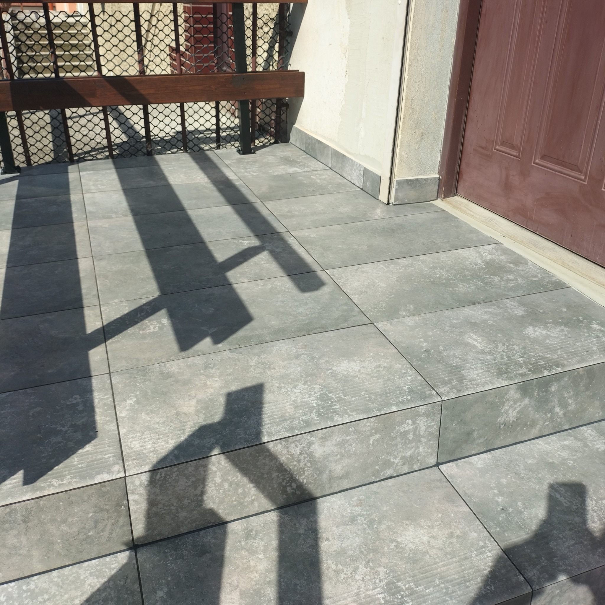 Szare płytki podłogowe na balkonie, widoczne cienie barierki, brązowe drzwi wejściowe w tle.