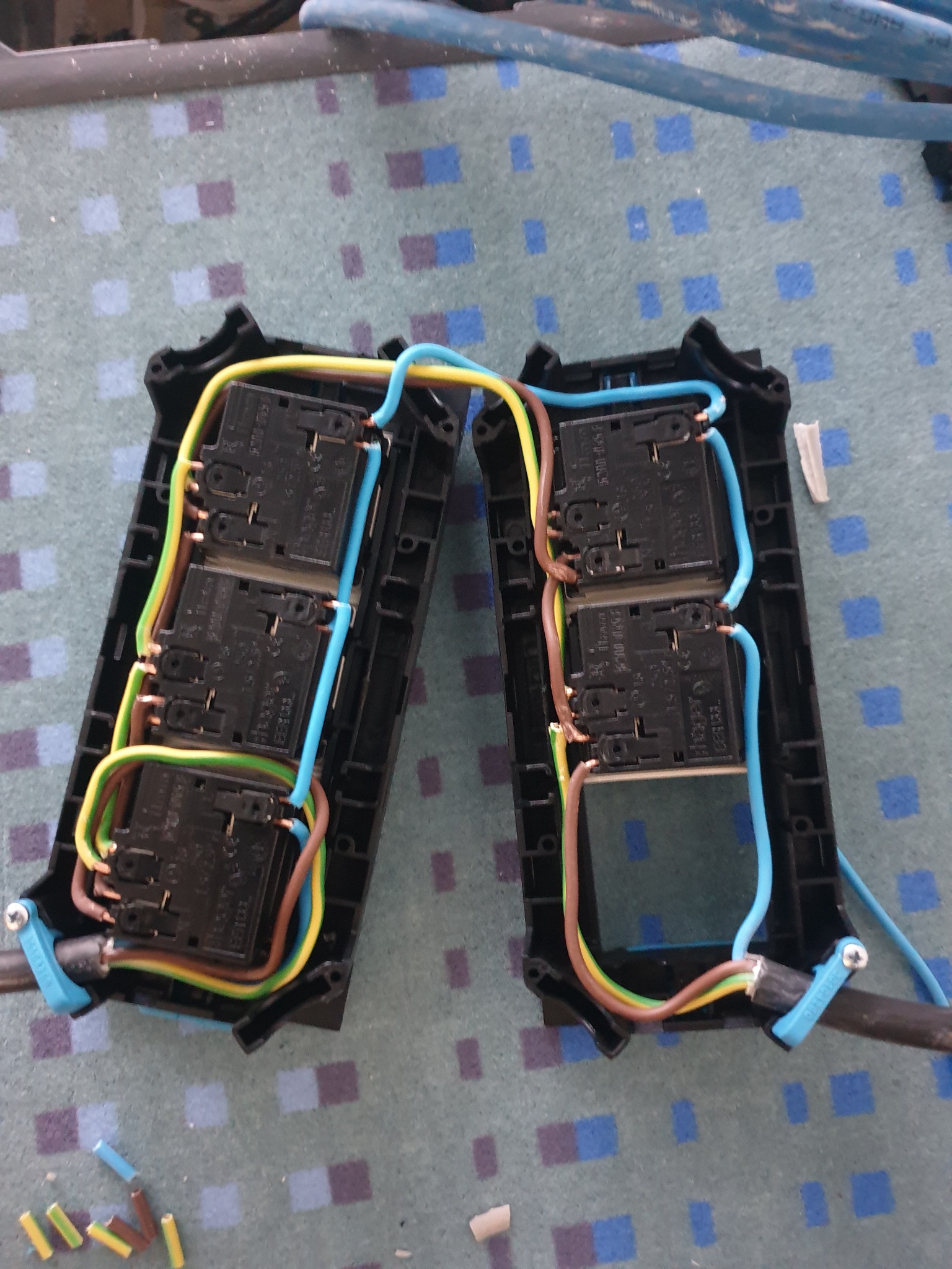 Dwa czarne rozdzielacze elektryczne z podłączonymi przewodami (niebieski, brązowy, żółto-zielony) na dywanie w niebiesko-szarą kratę, widoczne fragmenty kabli i końcówki przewodów.