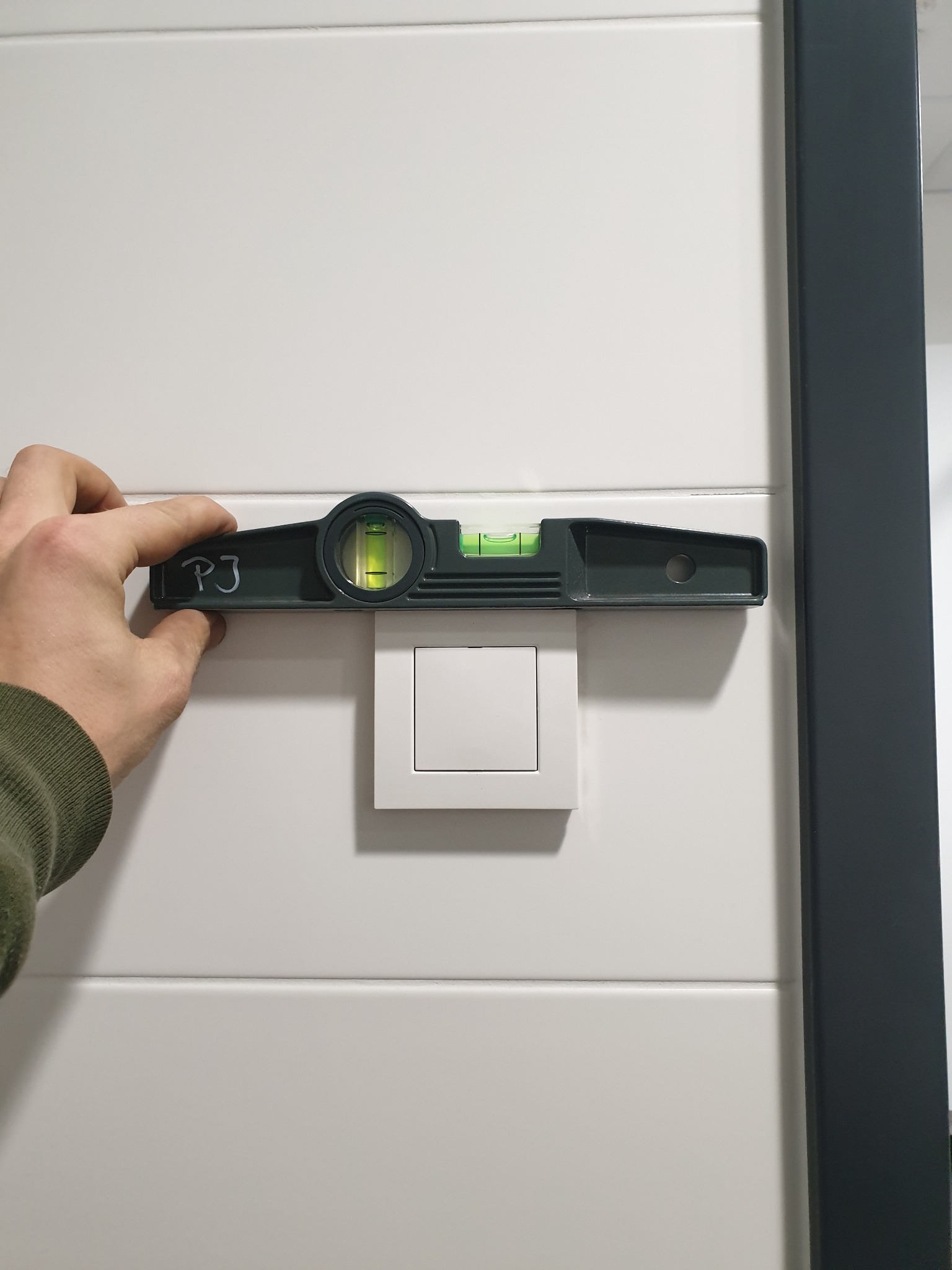 Poziomica z napisem 'PJ' przykładana do białego włącznika światła na białej ścianie z szarym obramowaniem.