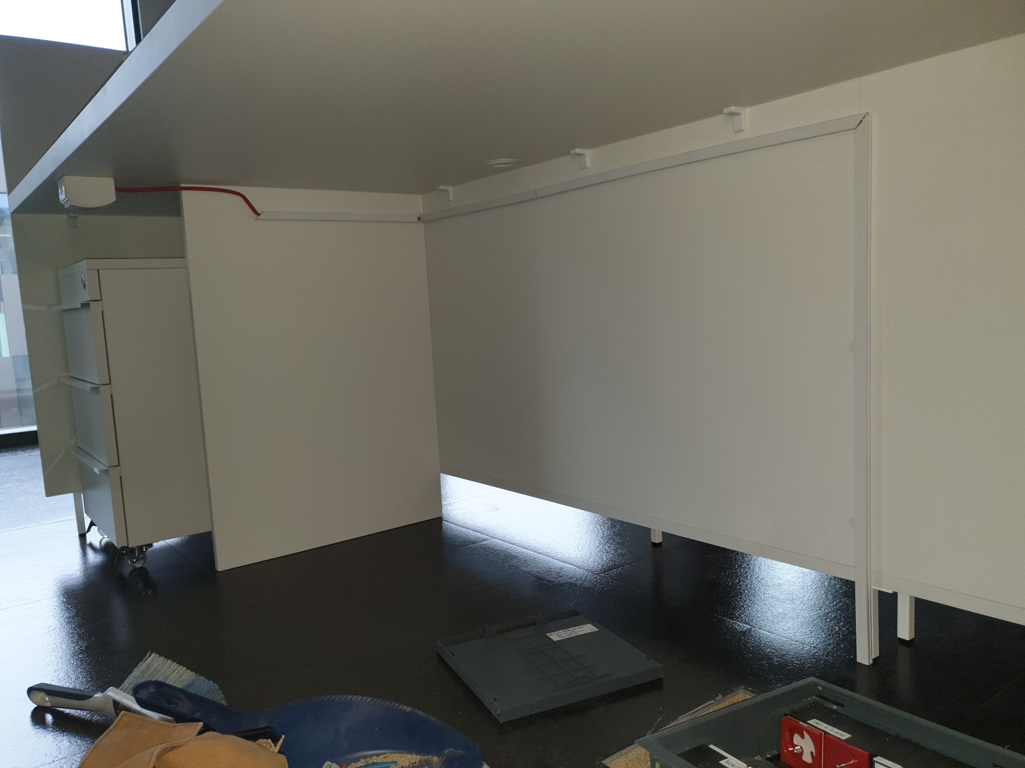 Widok spod biurka z widocznymi przewodami elektrycznymi, puszką instalacyjną i narzędziami sprzątającymi na podłodze.