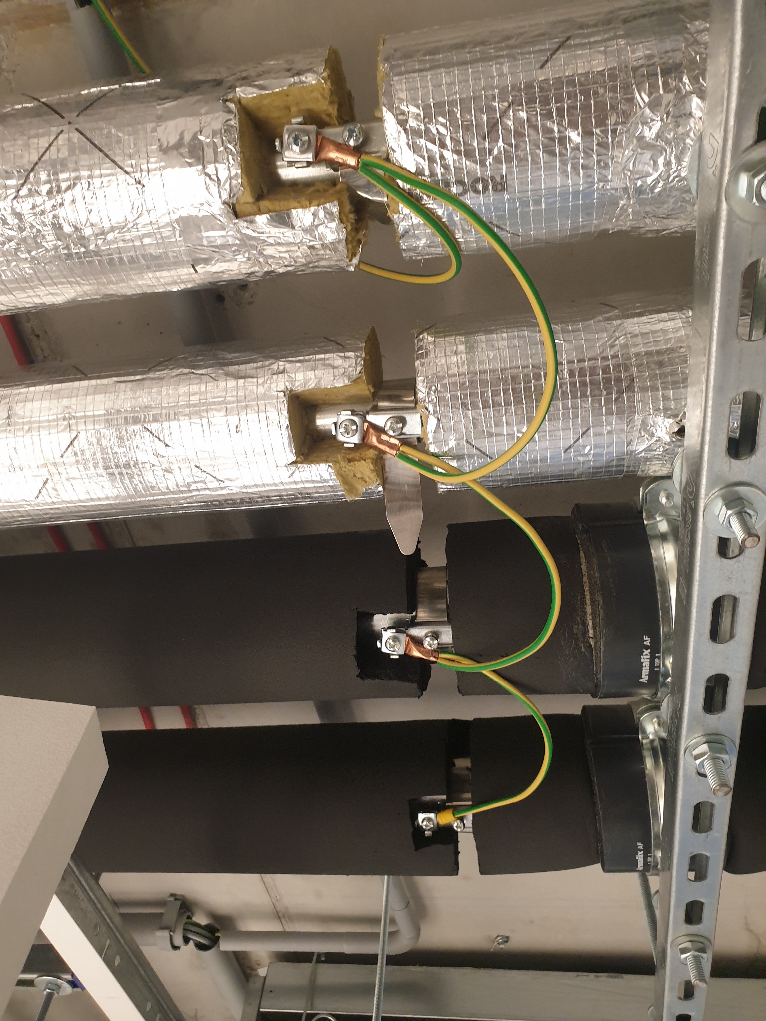 Uziemienie rur wentylacyjnych i instalacyjnych z użyciem przewodów żółto-zielonych i metalowych obejm, widoczne izolacje rur i konstrukcja wsporcza z metalu.