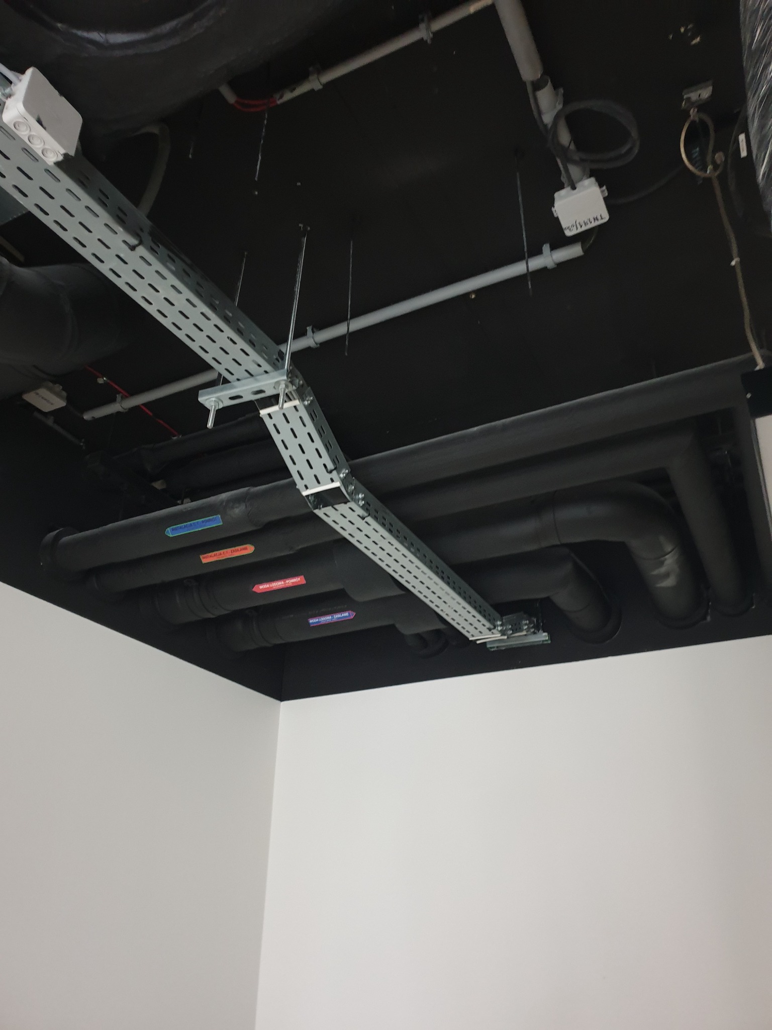 Widok instalacji pod sufitem: koryta kablowe z perforowanej blachy, czarne rury z izolacją i oznaczeniami, biała puszka elektryczna, czarny sufit i białe ściany.