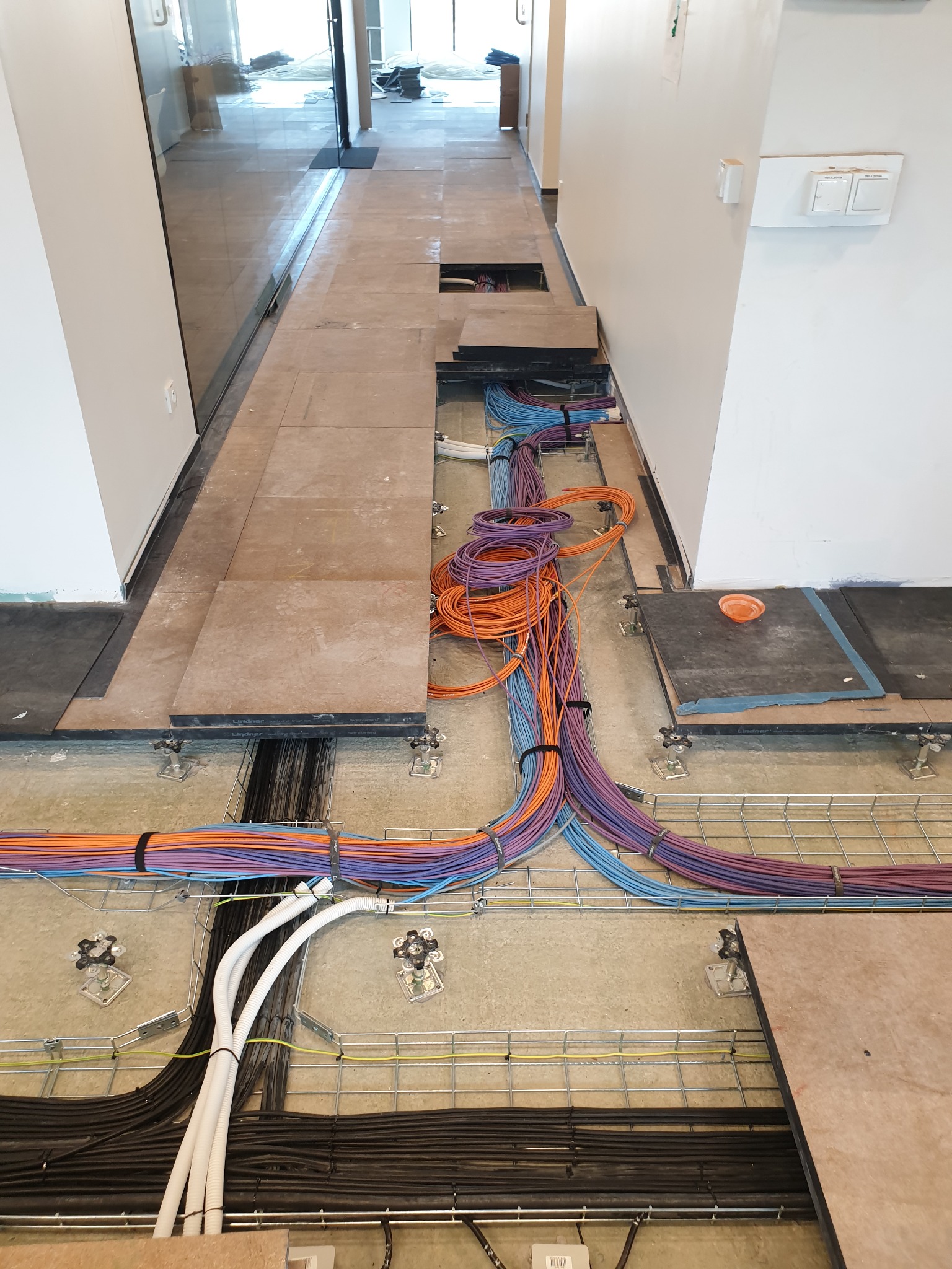 Otwarta podłoga podniesiona z widocznymi kablami elektrycznymi w korytkach, różnokolorowe przewody ułożone w pętle, fragment płyty podłogowej z pomarańczowym kubkiem.