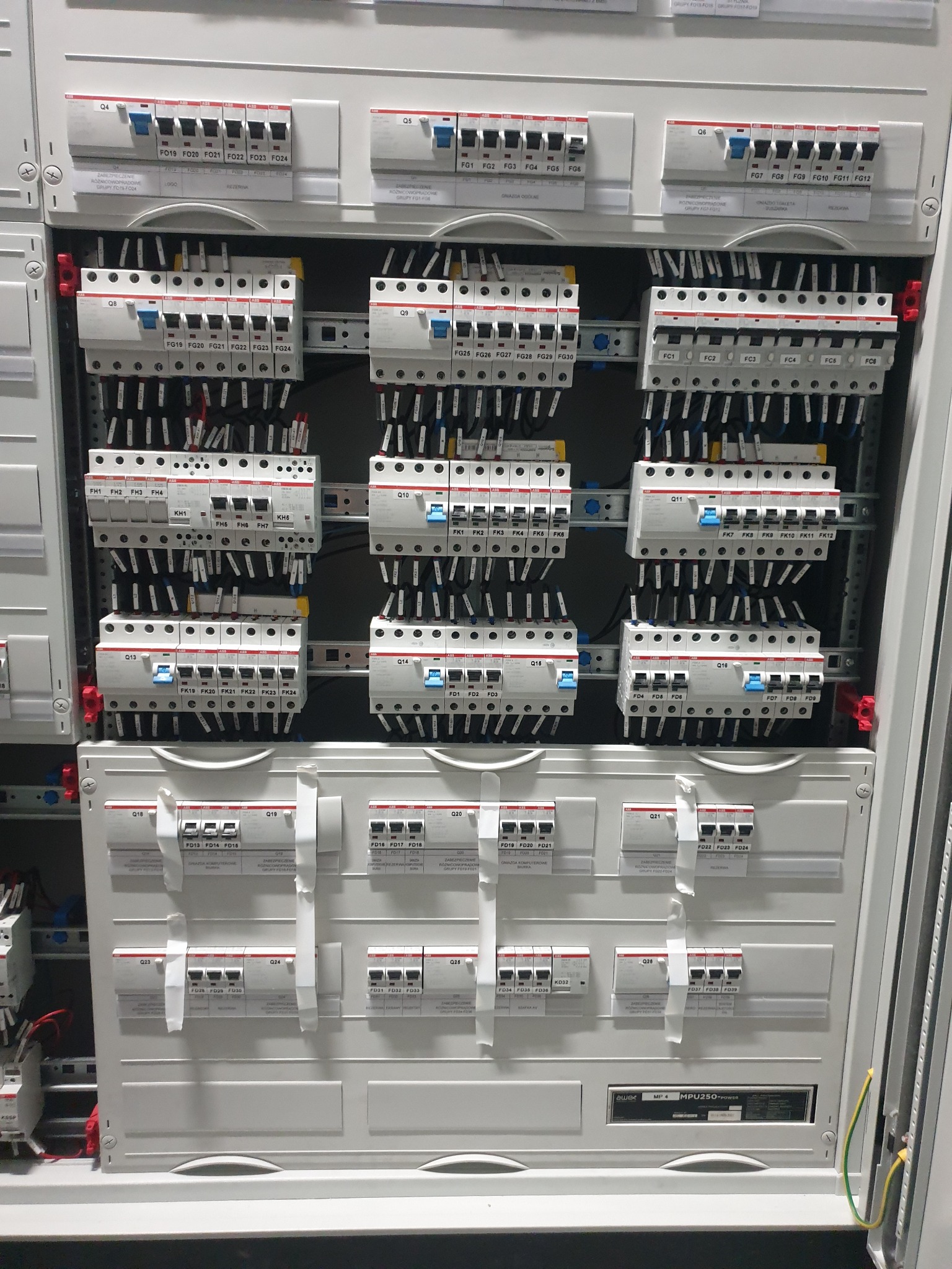 Rozbudowana, szara szafa elektryczna z licznymi bezpiecznikami, wyłącznikami różnicowoprądowymi i przewodami, widok z bliska, ukazujący skomplikowaną instalację elektryczną.