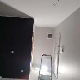 Wnętrze w trakcie remontu: ściana pomalowana na czarno kontrastuje z szarą ścianą, widoczna drabina, zabezpieczone drzwi folią, wystające kable oświetleniowe z sufitu.