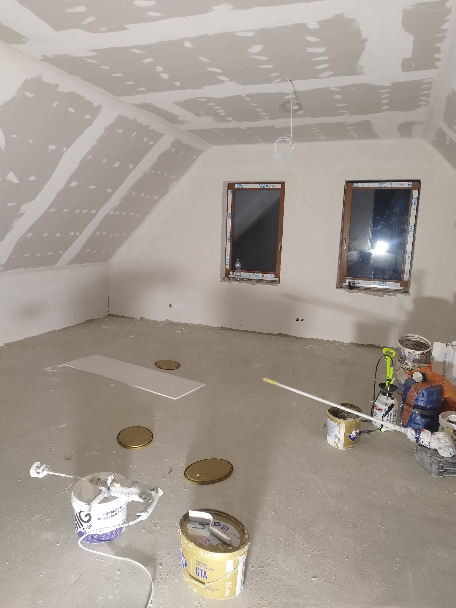 Poddasze w trakcie prac wykończeniowych, widoczne płyty gipsowo-kartonowe na skosach i suficie, zaszpachlowane łączenia, dwa okna z brązowymi ramami, na podłodze narzędzia i puszki z masą szpachlową.