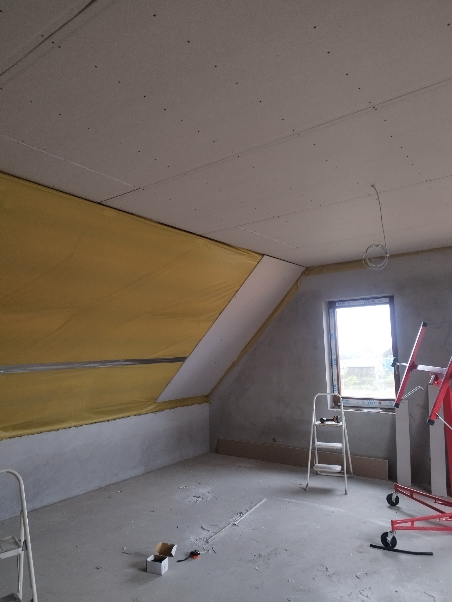 Wnętrze poddasza w trakcie adaptacji: ściany przygotowane do wykończenia, częściowo ocieplone, zamontowane płyty gipsowo-kartonowe na suficie, drabina i podnośnik do płyt w pomieszczeniu.