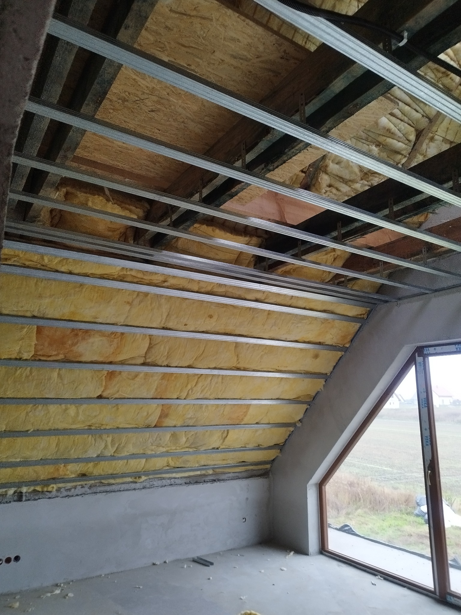 Montaż izolacji z wełny mineralnej w poddaszu, widoczne profile metalowe konstrukcji sufitu podwieszanego, okno w szczycie dachu z widokiem na zewnątrz.