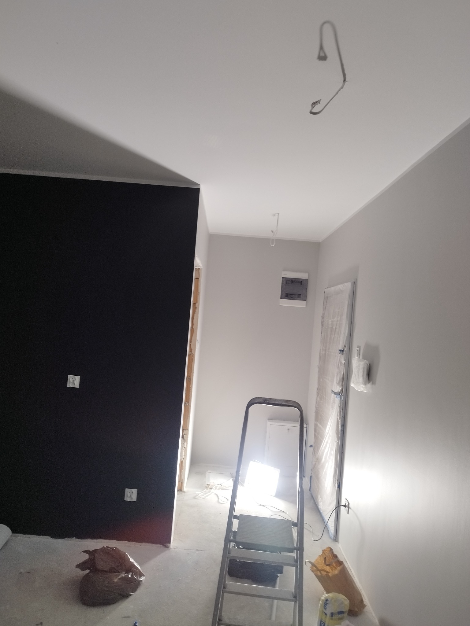 Wnętrze w trakcie remontu: ściana pomalowana na czarno kontrastuje z szarą ścianą, widoczna drabina, zabezpieczone drzwi folią, wystające kable oświetleniowe z sufitu.