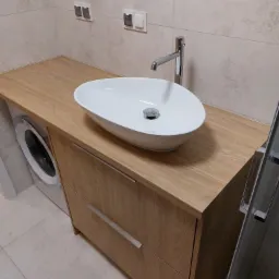 Łazienka z umywalką nablatową w kształcie łzy, zamontowaną na drewnianym blacie z szufladami, obok pralki.