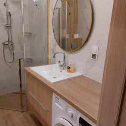 Nowoczesna łazienka z kabiną prysznicową w złotej ramie, okrągłym lustrem z ramą w kolorze złota, umywalką nablatową na szafce z blatem imitującym drewno, pralką pod blatem i marmurowymi płytkami...